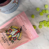 Pink package of Sour Apple Dots with a can of hot cocoa 
