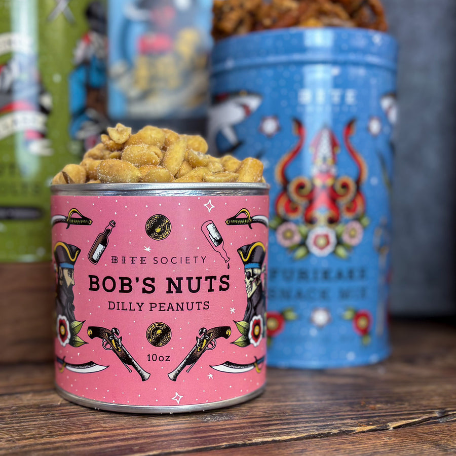 Can of 'Bob's Nuts' peanuts with a pink label featuring illustrations, on a wooden surface with other colorful tins in the background.