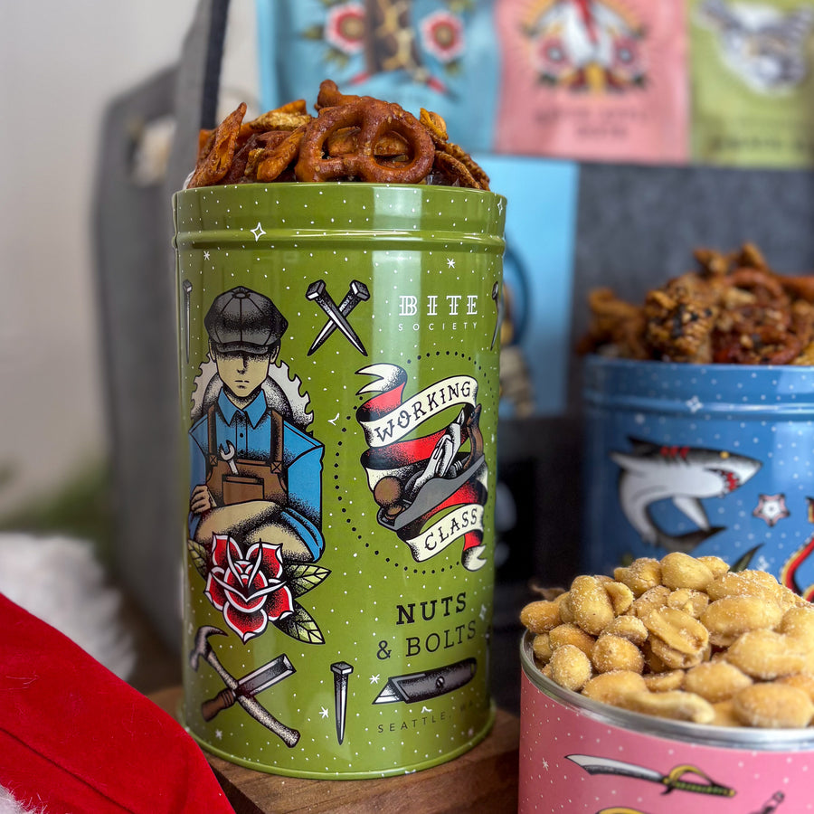 Green tin labeled 'Working Class Nuts & Bolts' with snacks on a wooden surface.