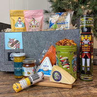 The Bite Before Christmas Gift Basket with assorted snacks and cheeses on a wooden surface with a decorative nutcracker cookie tin and holiday themed candy bags.