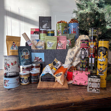 12 Days of Christmas Holiday Gift Basket with tins of cookies, cocoa, chips, and snacks. Along with bags of candy and a cooler of charcuterie and cheeses.
