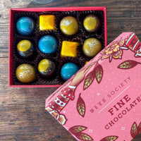 Box of colorful chocolates with a pink 'Bite Society' chocolate box on a wooden surface