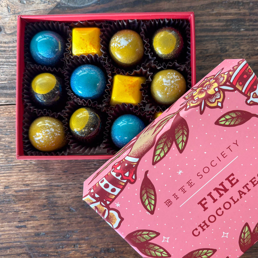 Box of colorful chocolates with a pink 'Bite Society' chocolate box on a wooden surface